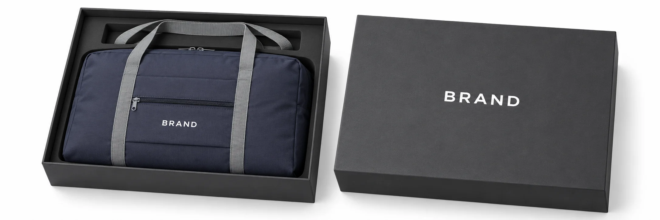 Navy gym duffel with gray handles and white lettering arranged inside a charcoal presentation box beside its lid.